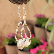 Load image into Gallery viewer, Silver twisted knot dangle earrings in a metal post.  Pink and purple flowers in planted pots in the background.  
