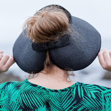 Load image into Gallery viewer, Model is wearing a black visor hat with hair in a bun. She has on a navy and green leaf shirt. Background includes rocks and ocean