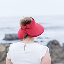 Load image into Gallery viewer, Model is wearing a red bow visor with brown hair in a bun, She has on a wihte shirt. There are rocks and ocean in the background.