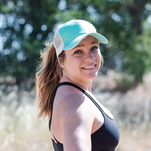 Model has brown hair and is wearing a blue cap.  Her hair is in a  ponytail.  She is wearing a black tank top.  Background is trees. 