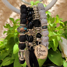 Load image into Gallery viewer, Black Tasseled Beaded Layering Bracelet hanging on white metal hook with green leaves in the background.