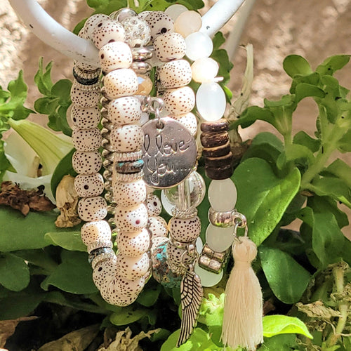 Cream Tasseled Beaded Layering Bracelet hanging on white metal hook with green leaves in the background. 