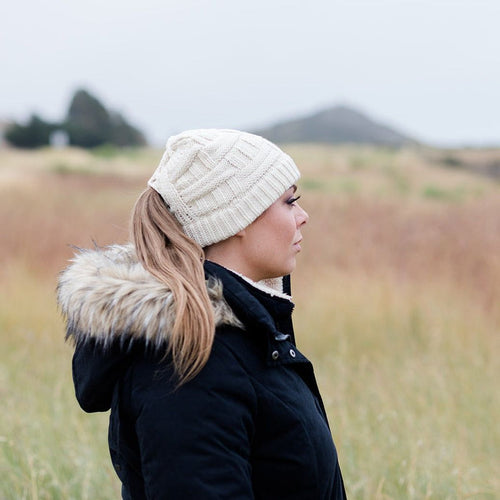 Cream Ponytail & Messy Bun Knit Beanie side view on model with brown hair in a ponytail and a black jacket.  Grass and mountains and bushes in the background 