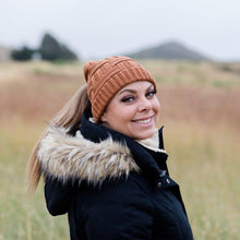 Load image into Gallery viewer, Brown Ponytail & Messy Bun Knit Beanie front view on model with brown hair in a ponytail and a black jacket. Grass and mountains and bushes in the background