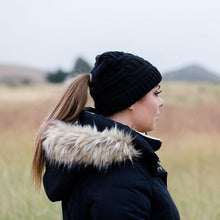 Load image into Gallery viewer, Black Ponytail & Messy Bun Knit Beanie side view on model with brown hair in a ponytail and a black jacket. Grass and mountains in the background