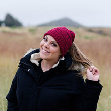 Load image into Gallery viewer, Burgundy Ponytail & Messy Bun Knit Beanie front view on model with brown hair in a ponytail and a black jacket. Grass and mountains and bushes in the background