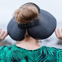 Load image into Gallery viewer, Model is wearing a black visor hat with hair in a bun.  She has on a navy and green leaf shirt.  Background includes rocks and ocean
