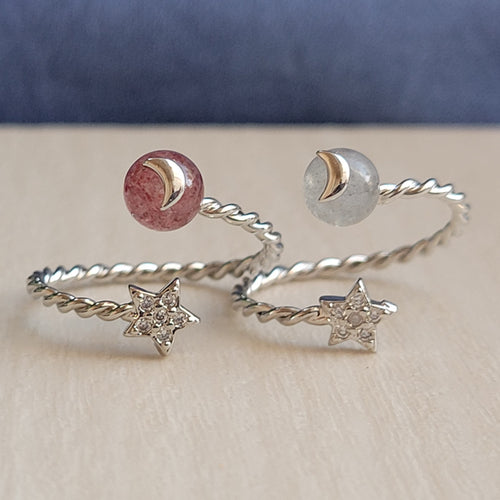 Picture of strawberry stone adjustable ring on the left and moonstone adjustable ring on the right. Rings are sitting on top of a tan surface with blue in the background.