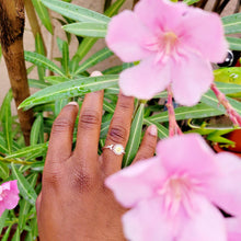 Load image into Gallery viewer, Picture of adjustable daisy ring on right ring finger with pink flowers and green stems in the background 
