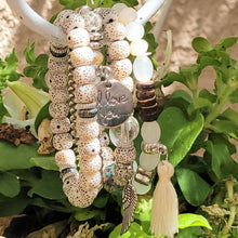 Load image into Gallery viewer, Cream Tasseled Beaded Layering Bracelet hanging on white metal hook with green leaves in the background. 
