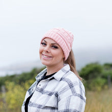 Load image into Gallery viewer, Pink Ponytail &amp; Messy Bun Knit Beanie front view on model with brown hair in a ponytail and a white flannel shirt with black stripes and a solid shirt under. Grass and bushes in the background
