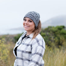 Load image into Gallery viewer, Black and White Ponytail &amp; Messy Bun Knit Beanie front view on model with brown hair in a ponytail and a white flannel shirt with black stripes and a solid shirt under. Grass, bushes, and mountains in the background
