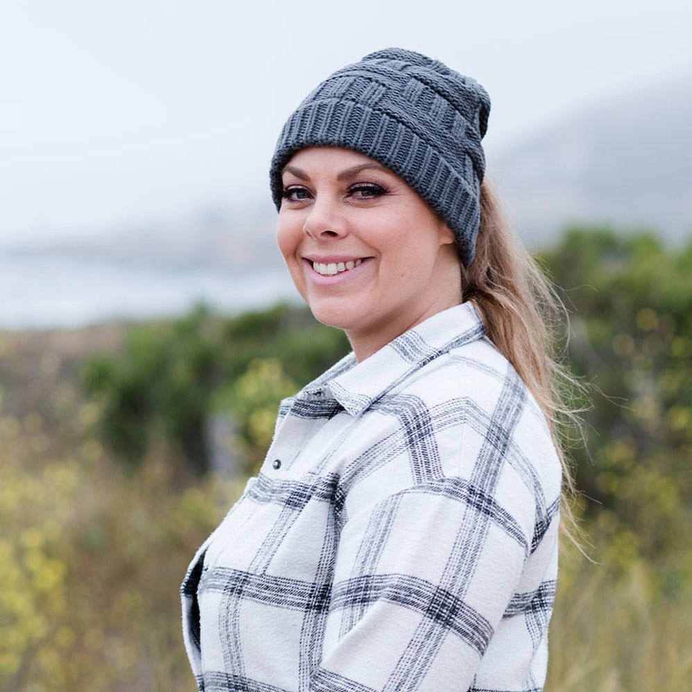 Grey Ponytail & Messy Bun Knit Beanie front view on model with brown hair in a ponytail and a white flannel shirt with black stripes and a solid shirt under. Grass, bushes, and mountains in the background