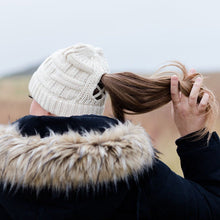 Load image into Gallery viewer, Cream Ponytail &amp; Messy Bun Knit Beanie back view on model with brown hair in a ponytail and a black jacket.  Grass and mountains in the background.  Model is twirling her hair. 
